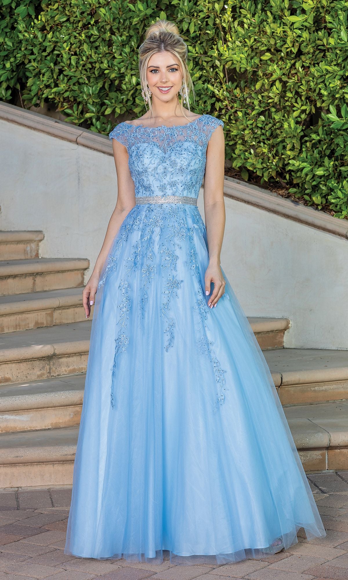 A woman stands on outdoor steps wearing the Corset-Back Illusion-Sweetheart Long Ball Gown, a light blue, floor-length lace dress with cap sleeves and a sheer neckline. Green foliage and a white wall create an elegant formal backdrop.